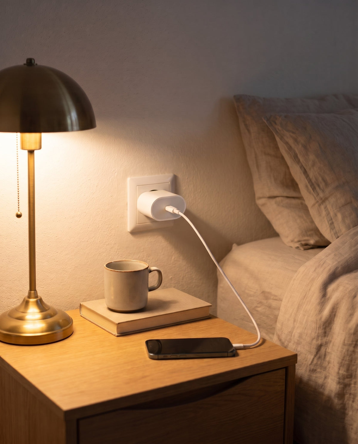 Adaptador de 20 W de Apple enchufado en la pared de un dormitorio, cargando un iPhone en la mesilla